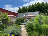 A walled garden terrace with a gravel path, green plants, patio umbrellas, a small table and chairs, a corrugated metal roof, and a red building in the background under a blue sky.