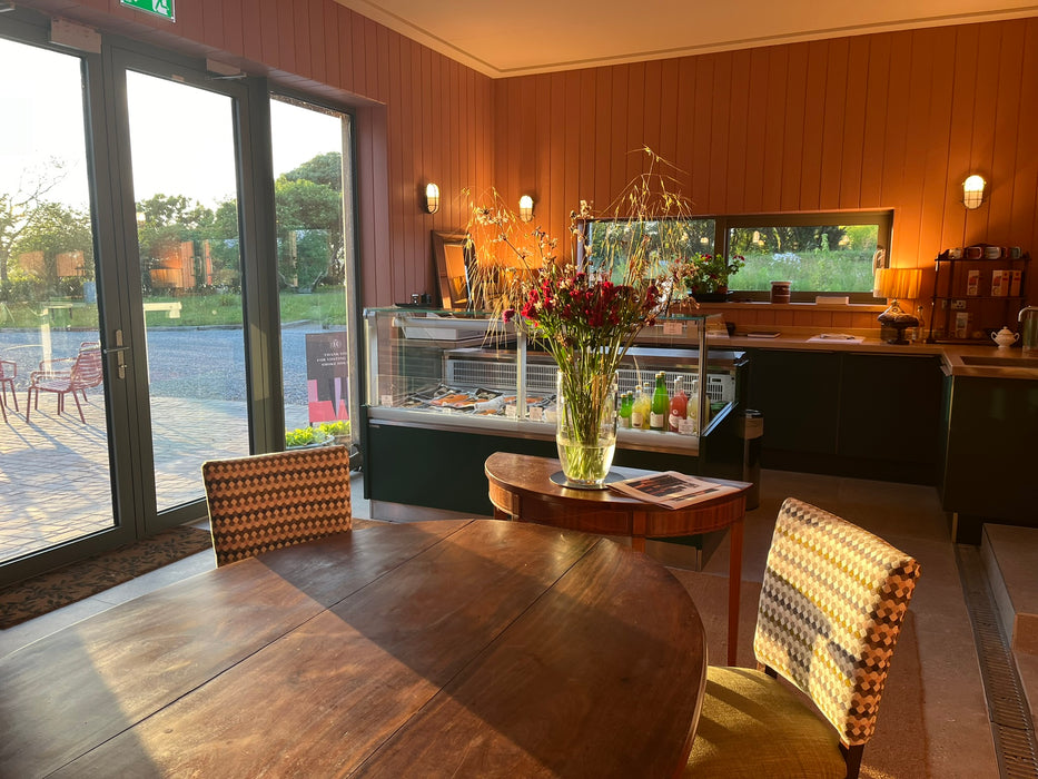 A warm, tasting room shop and dining area with wood-paneled walls, a wooden table and chairs in the foreground, a glass display case, and large windows looking out onto a courtyard and greenery.
