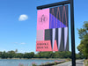 A vertical rectangular outdoor sign for Smoke House featuring a pink, purple, gray, and black geometric design with a stylized H logo and the words 'SMOKE HOUSE' in black text. The sign is mounted on a black metal pole outdoors near Cork harbour estuary and trees under a blue sky.