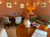 A cosy and comfortable interior room with wood-paneled walls, framed artwork, wall sconces, a dining table with smart upholstered chairs, a sofa with cushions, and a round wooden table in the foreground holding a white pitcher filled with red flowers, books, and an open notebook.