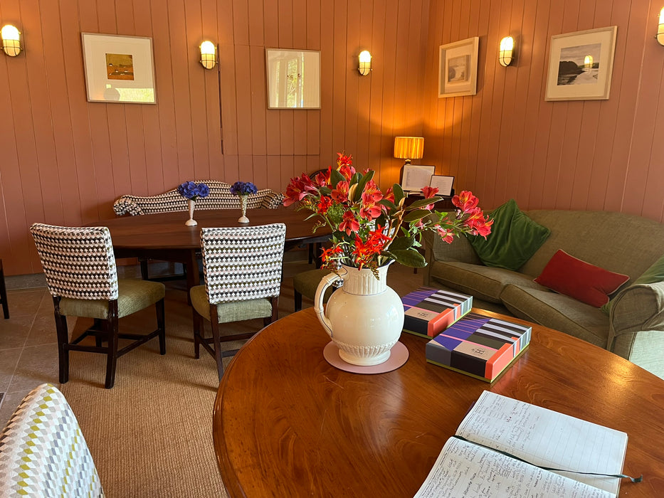 A cosy and comfortable interior room with wood-paneled walls, framed artwork, wall sconces, a dining table with smart upholstered chairs, a sofa with cushions, and a round wooden table in the foreground holding a white pitcher filled with red flowers, books, and an open notebook.